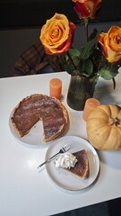 Homemade pumpkin pie with whipped cream slice served on a white table beside flowers and candles. The image reflects the concept of cozy autumn atmosphere and festive dessert tradition