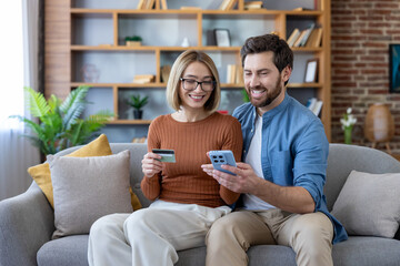 Happy young couple relaxing on a couch at home, collaborating on online shopping, holding a smartphone and credit card while making a secure e-commerce payment
