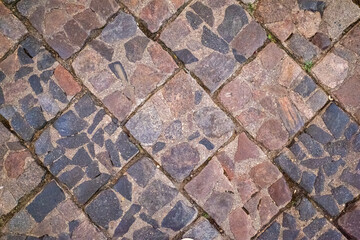 Overhead view of a cobblestone path, featuring small, irregular stones in square patterns with visible cracks and sparse grass.