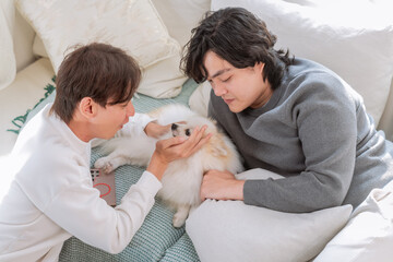 "LGBTQ male couple shares intimate bonding moments with beloved white Pomeranian pet dog companion on comfortable living room sofa, celebrating loving relationship precious family time together home