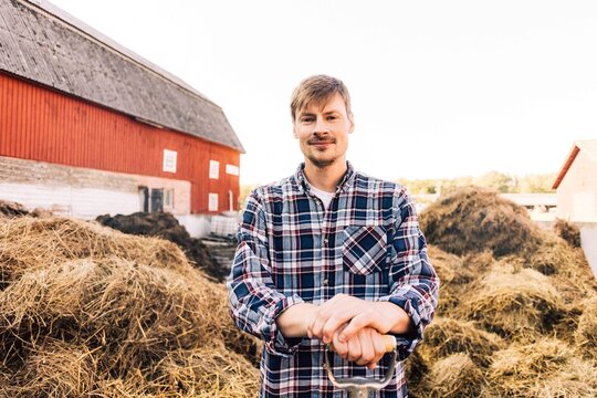 Portrait of confident farmer in plaid shirt posing against stack of hay at barn