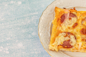 Close-up of a homemade salami pizza slice with melted cheese and crispy crust served on a decorative porcelain plate, placed on a light blue textured background in soft natural light.