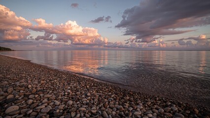 A peaceful lakeside scene with clouds reflecting on the water and a pebble shore at sunset.