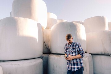 Male farmer using smart phone while leaning on hay stack in farm