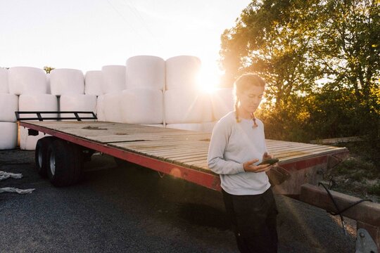 Female farmer using smart phone leaning on trailer in farm at sunset