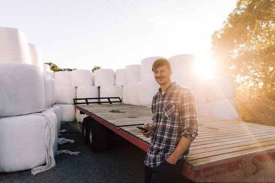 Portrait of smiling male farmer leaning on trailer holding smart phone