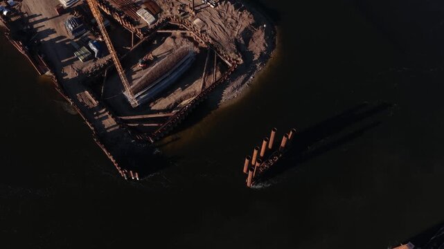 Aerial view of bridge foundation construction on a river, featuring heavy steel sheet piling, concrete reinforcement, and a tall tower crane on a cofferdam platform.