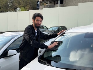 A young man smiling while carefully hand-washing or detailing the windshield and roof of a car with a black microfiber cloth. This image symbolizes car care, detailing service, and DIY maintenance.