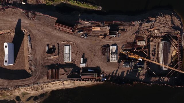 Vertical aerial image of a bridge foundation construction site on a riverbank, showing rebar, formwork, steel piles, and machinery within a sheet pile cofferdam.