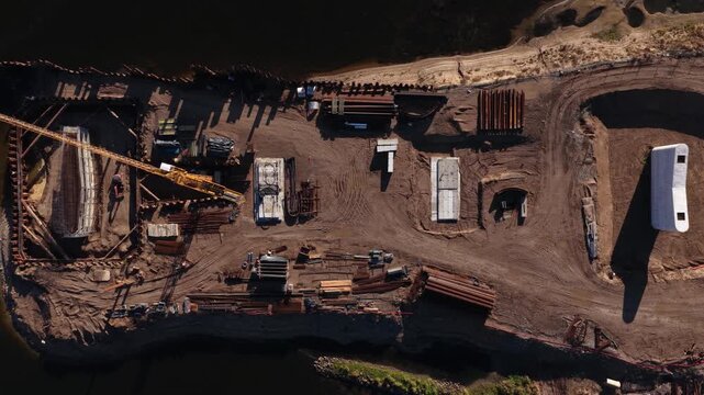 Vertical aerial image of a bridge foundation construction site on a riverbank, showing rebar, formwork, steel piles, and machinery within a sheet pile cofferdam.