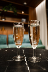 Two flutes of sparkling wine stand on black marble in Mellieha, Malta. Warm indoor light, teal seating, and wooden shelves appear soft as glossy reflections add a celebratory mood.