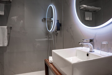Contemporary hotel bathroom in Mellieha, Malta shows a white vessel sink, chrome faucet, wooden vanity, glass, towel, and two backlit mirrors against gray marble look tiles.