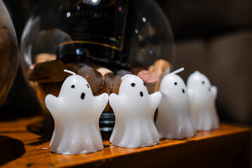Obraz premium Helloween vibes with four small white ghost candles sit on a polished wooden surface. A glass jar of wine corks and a dark wine label blur behind. Warm low light and shallow depth of field.