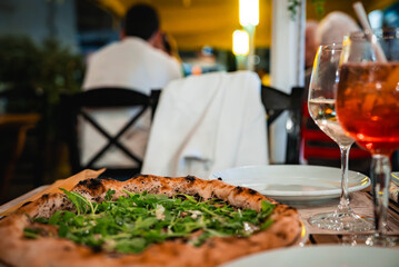 Wood fired pizza with arugula and two spritz sit on a wooden table in Valletta, Malta. Diners sit under yellow canopies and string lights on a narrow street at evening.