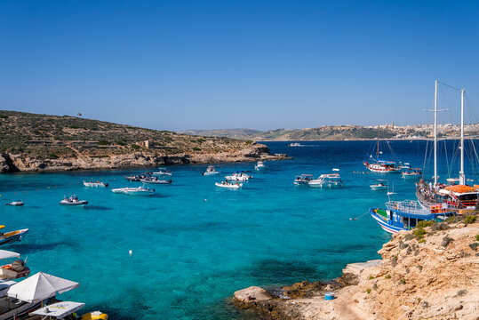 Turquoise shallows at Blue Lagoon, Comino, Malta show boats and yachts at anchor near limestone shores, swimmers and day trippers gather under clear summer light.
