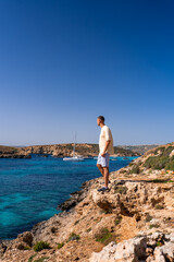 A solitary traveler stands on a limestone cliff, viewing Blue Lagoon in Comino, Malta. A white sailboat rests in a sheltered bay. Bright midday light, vertical frame.
