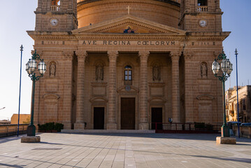 Grand sandstone facade of the Mellieha Parish Church in Mellieha, Malta, with portico, twin clock features, three wooden doors, green lamps, and long daylight shadows. © Aerial Film Studio