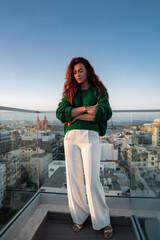 A woman in green sweater and white trousers stands on a glass terrace in Mellieha, Malta, facing a...