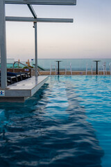 A rooftop infinity pool faces a glass balustrade with stools and tables in Mellieha Malta at dusk, with views toward Comino and Gozo, chaise lounges, and a lifebuoy.
