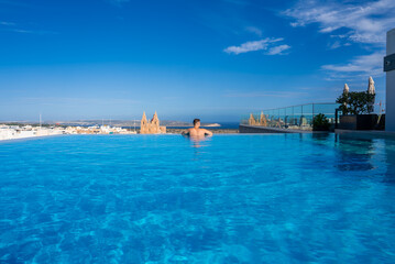 Naklejka premium A rooftop infinity pool in Mellieha, Malta, with one swimmer gazing toward the Mediterranean Sea, Gothic spires of the Sanctuary rising above town in bright midday light.