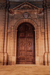 Fototapeta premium An ornate arched wooden door within carved limestone stands in Mellieha, Malta, evening lights warm the Baroque details, crest and pediment draw focus, stately mood