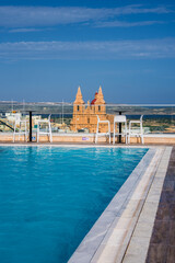A rooftop infinity pool with ladders and glass barrier faces the Sanctuary of Our Lady of Mellieha in Mellieha, Malta, with Comino and Gozo visible in bright midday light.