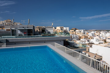 A rooftop infinity pool on a boutique hotel terrace in Mellieha, Malta, frames limestone buildings and blue water views toward Comino and Gozo in bright midday light.