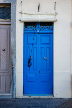 A cobalt blue door marked 82 stands in a narrow Marsaxlokk street in Malta, with twin knockers, iron transom, cream awning, and a beige door beside it in soft daylight.
