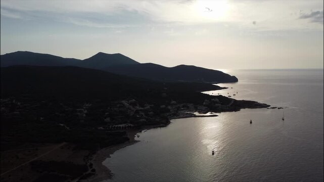Aerial view of Barcaggio village and coastline at sunset, northern Corsica, France. Calm sea and boats under golden evening light.