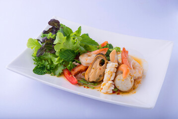 Seafood salad, a Thai dish. Shrimp, squid, mussels, and scallops, all boiled, with small tomato pieces, mint leaves, and salad greens. Placed on a white plate, white background.