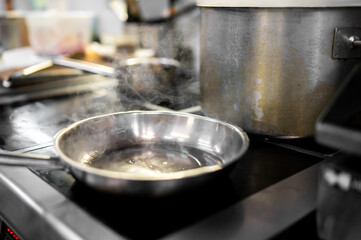 A stainless steel frying pan with oil heats on an electric stovetop, steam rising from the hot surface in a professional or home kitchen setting