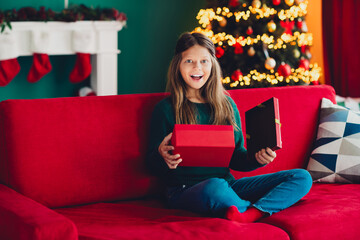 Obraz premium Funny festive surprise little girl opens red gift on a cozy red sofa beside a sparkling Christmas tree at home during a warm holiday celebration