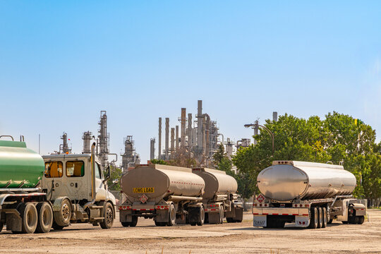 A fleet of large, silver tanker trucks is lined up for fueling with oil refinery in background.