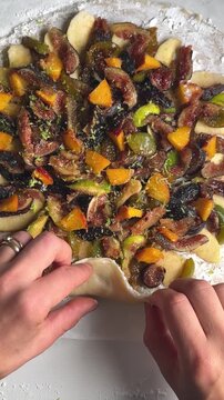 Close-up of a pastry chef folding the edges of fresh fruit tart dough filled with figs, peaches, pears and plums before baking.