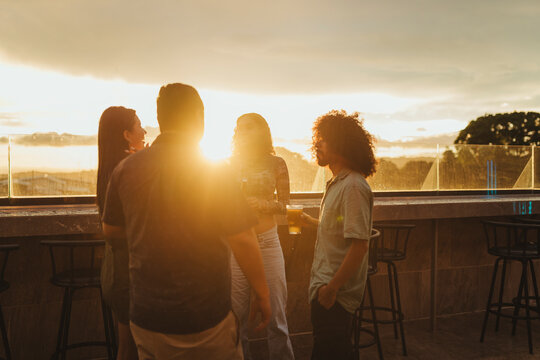 Group of young adults standing at a rooftop bar during sunset, enjoying drinks and socializing.  - Powered by Adobe