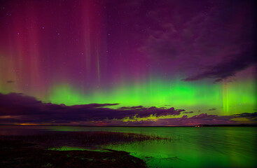 Brilliant aurora borealis in green and purple hues shines above a calm lake and cloudy night sky with faint stars.