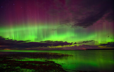 Vibrant aurora borealis in green and purple hues illuminating the night sky over a calm lake with reflections and clouds.