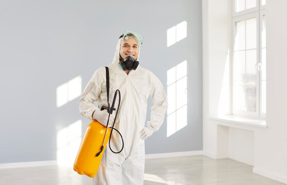 Portrait of man from disinfection service wearing protective white ppe suit and face mask looking at camera after disinfecting office or empty apartment with disinfectant spray. Protection concept.
