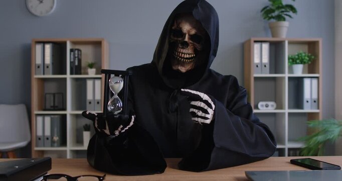 Terrible grim reaper, skeleton or devil in black costume posing at office desk, holding sand clock, demonstrating passage of time and that death is coming, looking scarying and horrible. 