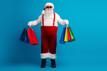 Santa with red suit and suspenders holds colorful shopping bags against a bright blue background ready for Christmas shopping and holiday celebration