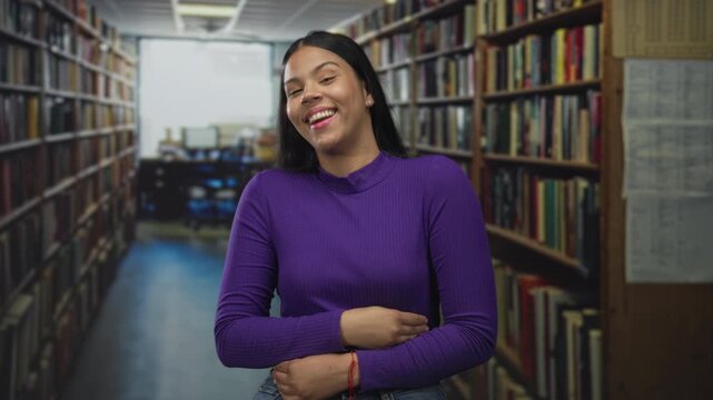 Woman laughs and holds her stomach amid tall library shelves in a quiet aisle wearing a purple sweater; happiness. - Powered by Adobe