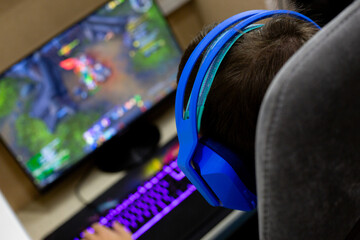 Back view of gamer wearing blue headphones playing online game with friends. Tilted camera angle shows gaming monitor with interface, RGB keyboard, home setup creating dynamic atmosphere