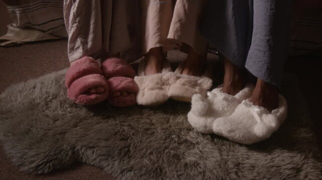 Medium shot of legs of anonymous multiethnic young women in pajamas and cozy plush slippers tapping rhythm on fur rug in bedroom, while enjoying music at slumber party