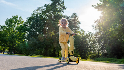 Scooter child. Girl riding scooter toddler. Yellow outdoor activity park road.