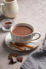 Hot chocolate in a cup on a light background with cinnamon, milk jug and napkin.