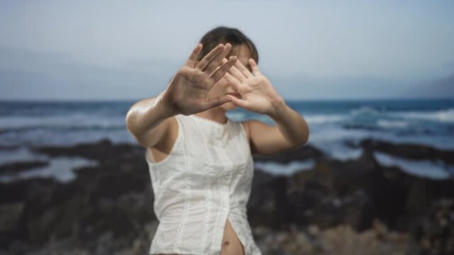 Young woman with hands raised to block camera at rocky seaside, white sleeveless blouse and palms forward; privacy solitude.