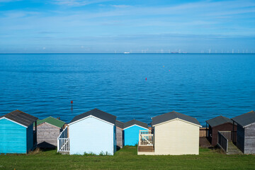 Wooden beach huts along a grass verge by the pebble beach in Tankerton, Whitstable in Kent. The sea and sky are a beautiful blue. There are wind turbines on the horizon.