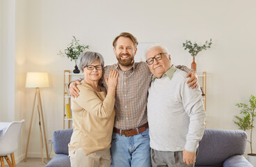 Portrait of happy millennial 35s man hugging his older parents, standing together in modern living room, posing for camera with positive expression. Family Day celebration, love and bonding concept