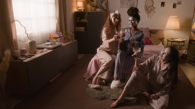 Full portrait of three happy diverse girlfriends, wearing cozy pajamas, sitting in bedroom with face masks, looking at camera, laughing, swaying to music, while chilling out together at home party