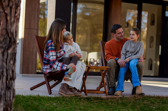 Happy family, including parents and two children, sitting together on chairs and a father's lap while relaxing and interacting comfortably on the patio of their modern home